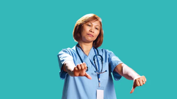 Close up of upset hospital worker showing thumbs down signs while at work, isolated over studio background. Dissatisfied asian healthcare expert doing frantic disapproval gestures