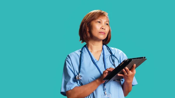 Licensed nurse wearing protective scrubs imputing medical data on tablet. Healthcare specialist happy at work, typing on digital screen, isolated over blue studio background
