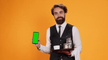 Genç garson kamerada yeşil ekran gösteriyor, elinde boş bir telif alanı görüntüsü olan akıllı telefon var. Dost canlısı zarif yemek çalışanı stüdyoda izole edilmiş boş ekran şablonuna işaret ediyor..