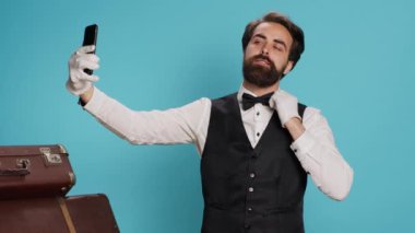 Stüdyoda, üniformalı kapıcı resimlerini çekerken akıllı telefonu kullanarak mavi arka planda aptalca davranır. Yetenekli otel kapıcısı iş üstündeyken aletleriyle resim çeker..
