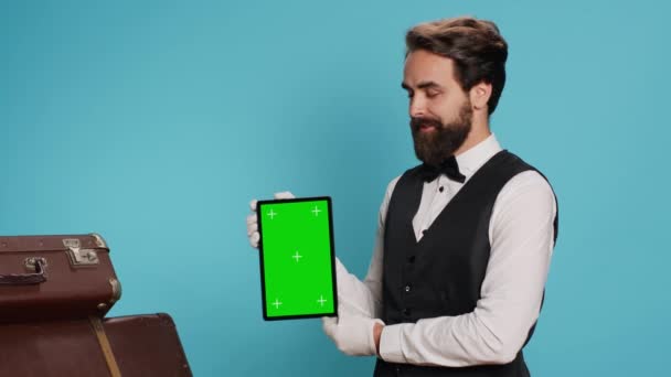 时尚的行李员在工作室里展示平板电脑 并展示空白的绿色显示屏 而他站在推车袋旁边 优雅的搬运工用豪华服务展示礼宾服务的协助 — 图库视频影像