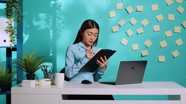 Determined hardworking asian employee cross-checking bureaucratic data between laptop screen and clipboard notes. Professional employee working at modern office desk over blue studio background