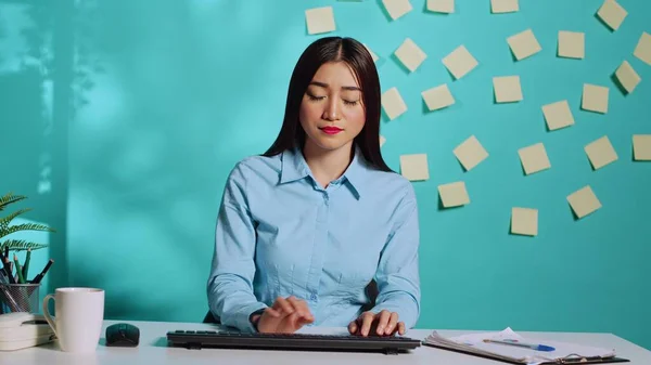 Focused asian office clerk typing on keyboard while receiving information from management during online videocall meeting. Employee at colourful modern workplace desk over blue studio background