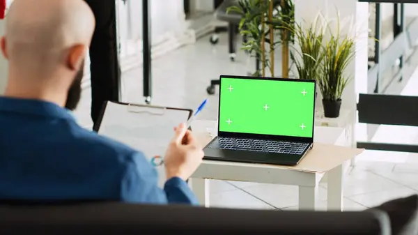 Businessman looks at greenscreen in office, using laptop to plan company strategy. Middle eastern man working on isolated copyspace display, entrepreneurship concept in coworking space.