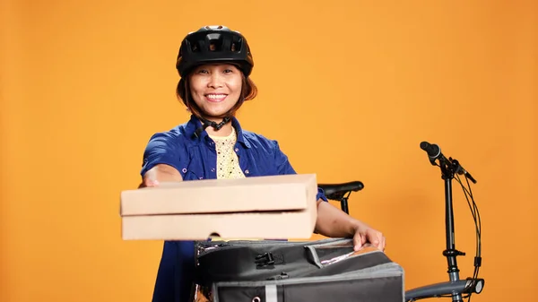 Friendly pizza delivery woman wearing bike gear bringing order to customer. Upbeat asian courier getting lunch boxes out of thermal food backpack, isolated over orange studio background