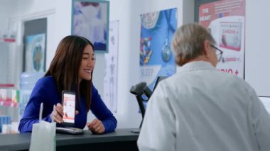 Eczane tezgahındaki müşteri, eczacının kalp atışı için reçete yazdığını akıllı telefondaki hapları düzenleyip satın almaya hazır olduğunu gösteriyor. Yaşlandırılmış sağlık uzmanı müşteri satışı için gerekli tedavi ürünü