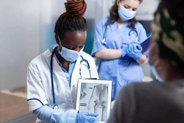 Bone doctor explaining clinical information on tablet to patient during orthopedic medical exam. Orthopedist in modern sterile hospital office wearing face mask and medical gloves