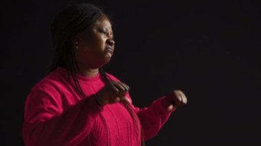 Siyah arkaplan modeline karşı durmak ellerini önünde tutarak onaylamadığını gösteriyor. Pembe kazaklı Afro-Amerikalı kadın karşı çıkmak için baş parmak hareketi kullanıyor. Yan görünüm.