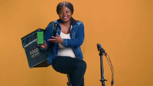非裔美国人信使手持手机 坐在自行车上展示绿色屏幕 在带有空白模拟模板的移动设备上展示创新交付应用程序的黑人妇女 — 图库视频影像