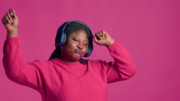 女性时尚博主使用无线耳机 享受最喜爱音乐的节拍 穿着粉色套头毛衣的漂亮黑人女子在与世隔绝的背景下轻声地听着好听的音乐 — 图库视频影像