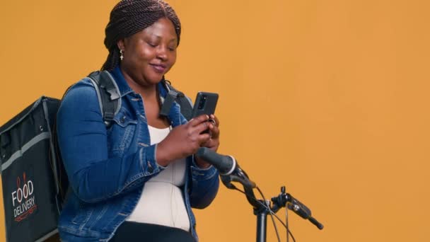 女性の自転車メッセンジャーは オンライン注文を待っている間 携帯電話上のメッセージを笑います 自転車でモバイルデバイスでブラウジングする熱狂的なアフリカ系アメリカ人宅配業者 ハンドヘルドショット — ストック動画