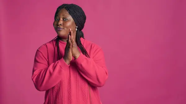 Glamorous female with african american ethnicity motioning to the side with left-hand. Stunning black woman with elegant style gesturing and applauding in front of pink color background.