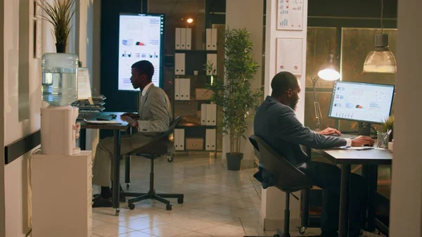 アフリカ系アメリカ人の従業員は 夜勤中に仕事を楽しみ 机の椅子でレースを開催しています オフィスで一晩滞在し 一緒に遊んでいるリラックスしたコワーキングビジネスマン — ストック写真