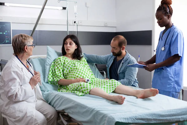 Diverse medical team discussing with pregnant woman before caesarean surgery in hospital ward. Patient with pregnancy lying in bed having contractions, being comforting by husband in maternity clinic