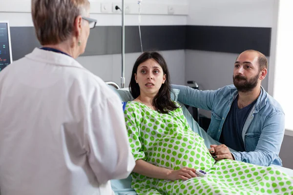 Couple discussing childbirth process with doctor in hospital ward, pregnant woman lying in bed preparing for medical surgery. Patient with pregnancy holding hands on belly being comforting by husband
