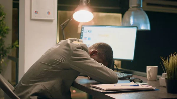 アフリカ系アメリカ人のコンピュータオペレーターが オフィスで一晩働きながら机の椅子で眠っている 仕事で昼寝をする疲れた従業員 未完成のタスクを持つワークスペースで一人で — ストック写真