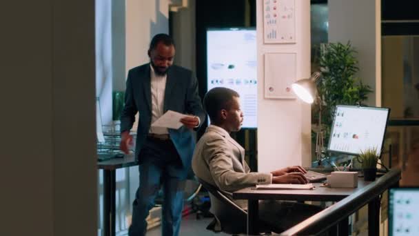 アフリカ系アメリカ人のスーパーバイザーが会計士に ナイトシフト中の近代的なオフィスでのビジネスプロジェクトの進捗状況を説明するように頼んだ コンピュータで作業する従業員を監督する経営幹部 — ストック動画