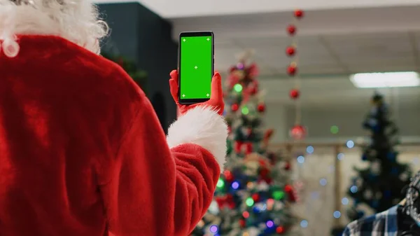 Supervisor in xmas ornate shopping mall wearing Santa Claus suit holding mockup phone, reading promotional Christmas offers for clients on green screen during winter holiday season, close up