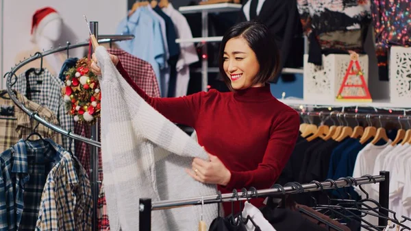 Asian customer browsing through rack of clothes in Christmas themed store, checking for fitting garment size. Woman in xmas adorn fashion boutique during winter holiday season