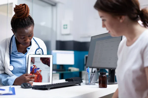 Cardiac specialist doctor explaining heart informations to patient on tablet at hospital desk. Cardiologist providing healthcare cardiac diagnosis to african american woman in clinical consultation