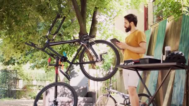 健康的高加索人在使用他的数码平板电脑进行指导的同时 还肩负着检查和修理自行车的多重任务 年轻而专注的男性骑自行车者浏览互联网以维护现代自行车 — 图库视频影像