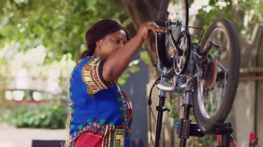 Hevesli siyahi bir kadının, bakım için bisiklet tekerindeki hasarı dikkatle incelerken çekilmiş portresi. Genç Afrikalı Amerikalı kadın bahçede bisikletini tamir ediyor, pedalları tamir ediyor..