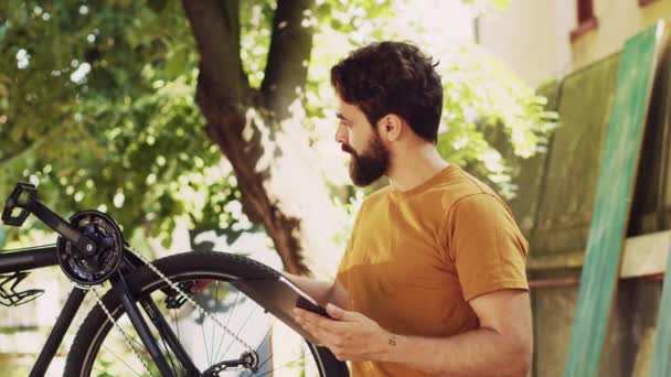 彼のデジタルタブレットを使用してガイダンスのための彼の自転車を調べ 修理することを決定したアクティブな男 ヤングスポーティ専用男性サイクリスト庭で自転車を維持するためにインターネットを閲覧 — ストック動画