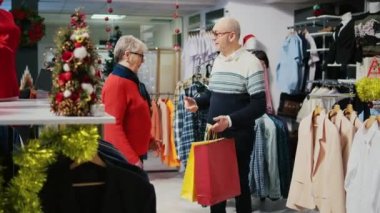 Elinde alışveriş poşetleri tutan yaşlı bir adam, şenlikli moda dükkanında dönen bir eş, Noel partisinde aile üyeleriyle paylaşmak için ideal hediyeler bulduktan sonra mutlu.