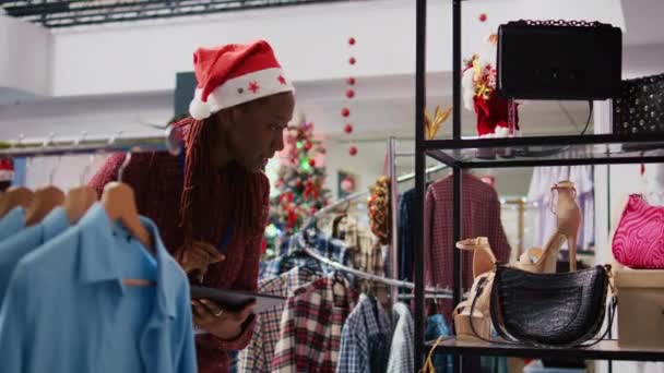 アフリカ系アメリカ人のスーパーバイザーは クリスマスのテーマのファッションブティックで服のラックをブラウズして ホリデーシーズンの販売中にオンラインストアでプロモーション価格を入力するタブレットを使用して — ストック動画