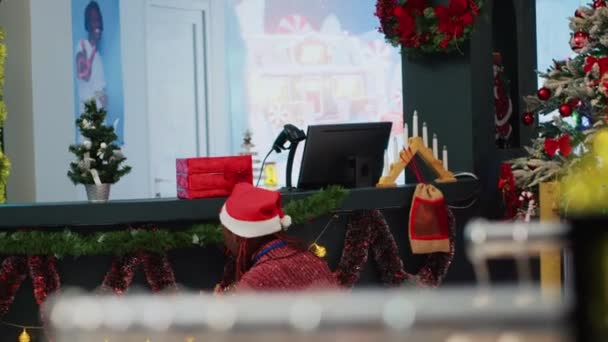 サンタの帽子をかぶったアフリカ系アメリカ人の従業員は お祝いのプロモーションイベントの前にクリスマスの装飾品店を飾ります 同僚が支援するファッションショップにXmasツリーをインストールする労働者 — ストック動画