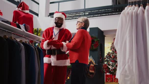 员工穿着圣诞老人的服装 向希望在圣诞装潢店为家人购买礼物的年长客户提供有关红色服装的材料和价格信息 — 图库视频影像