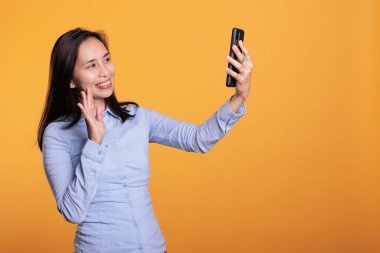 Video görüşmesi sırasında uzaktan kumandalı arkadaşıyla konuşan neşeli Asyalı bir kadın stüdyoda iş molası sırasında uzaktan tartışıyordu. Arama için telefon kullanarak sarı arkaplan üzerinde duran model