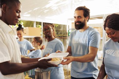 Friendly volunteers handing out free food to the hungry poor and needy individuals, outdoors. Underprivileged and homeless people receiving warm meals from the humanitarian aid team at a food drive. clipart