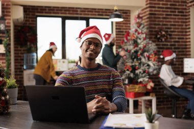 Gülümseyen Afro-Amerikan şirket çalışanı Noel Baba şapkası ve kışın örülmüş süveter giyerek ofis masasının önünde oturuyor. Noel 'de dekore edilmiş işyerinde kameraya bakan neşeli bir şirket çalışanı.