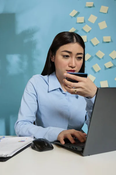 Corporate asian female employee using a laptop for Internet banking holding credit card. Businesswoman typing payment details sitting in the office, reviewing bank statement on secure website.