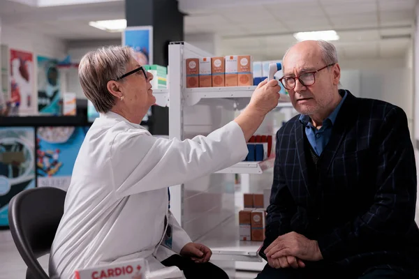 Pharmacist measuring old patient body temperature with thermometer gun, doing health check up and medical consultation. Pharmaceutical specialist testing senior man in glasses for fever in drugstore