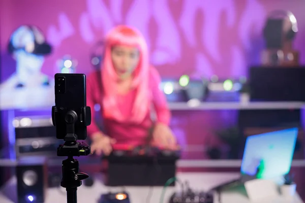 スマートフォンカメラでプロのミキサーコンソールを使用してエレクトロニックミュージックを行うピンクの髪を持つ女性 クラブの夜の間に歌をミキシングする笑顔アーティスト — ストック写真