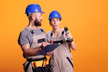 İnşaat işçileri not alıyor ve yenileme ölçümlerini kontrol ediyor, ellerinde matkap tabancası var. Kadın ve erkek tamirciler, sarı arka planda duran evrak ve dosyalara bakıyorlar..