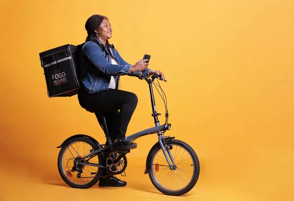 Restaurant employee carrying thermal takeaway backpack while delivering takeout food order to client, holding phone to check address on fast food app. Courier standing over yellow background in studio