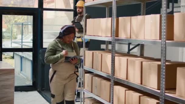 冷やされたアフリカ系アメリカ人の倉庫労働者は 段ボール箱のラベルをチェックしながら音楽を聞いている 貯蔵ハブの列を歩いている間 ヘアホンを身に着け 頭をぶつける笑顔の労働者 — ストック動画
