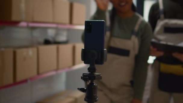 倉庫ロジスティクスコーディネーターや同僚が三脚に設置したスマートフォンを使ってインターン向けのトレーニングビデオを作成 従業員はフルフィルメントセンターで自分自身を撮影し トレーナーはボックスをシールする方法を示します — ストック動画