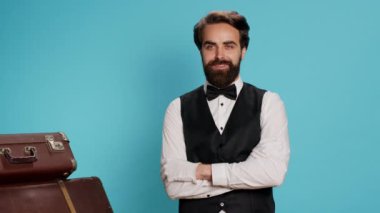 Mavi arka planda kameraya poz veren profesyonel otel kapıcısı bavulları taşımaya ve yolculara yardım etmeye hazırlanıyor. Bellboy olarak çalışan yetenekli bir işçi, turizm endüstrisi konsepti..