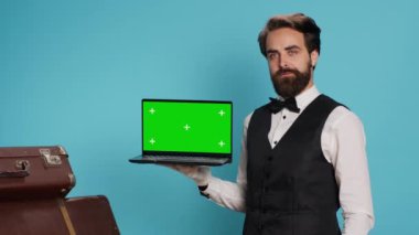 Klas komi, misafir valizinin yanında beyaz eldivenler giyerken stüdyoda dizüstü bilgisayarda yeşil ekran gösteriyor. Modern otelde çalışan lüks şık kapıcı, izole edilmiş bir model sunuyor..