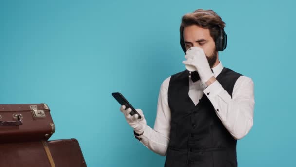 看门人发短信 在工作室里喝咖啡 欣赏耳机上的音乐 年轻的成年人 有优雅的酒店部门的工作信息和喝冷饮饮料 — 图库视频影像