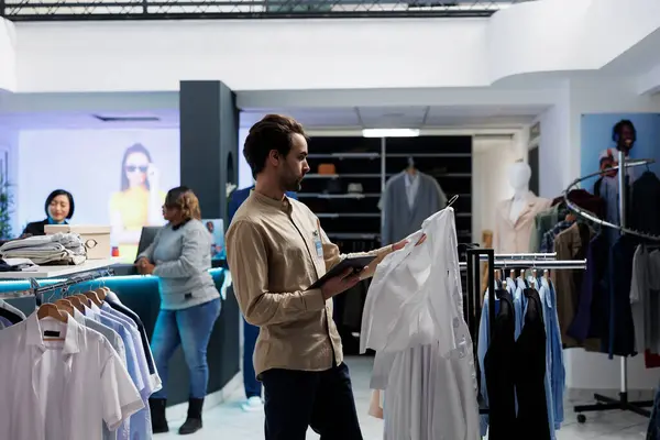 Clothing store employee checking shirt size and managing inventory using digital tablet. Fashion boutique manager holding apparel on hanger and examining merchandise in stock