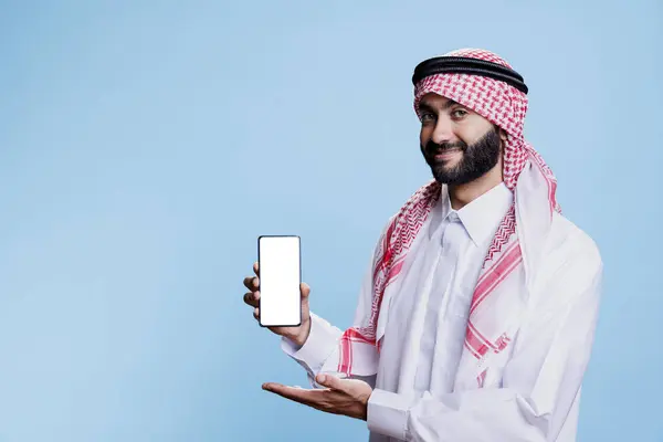 在传统的现场微笑的穆斯林男子给智能手机提供了空白的白色屏幕肖像 自信的Arab展示手机空白触摸屏和摄像头 — 图库照片