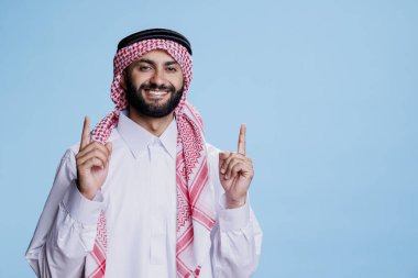 Geleneksel kültür kıyafetleri giyen gülümseyen Müslüman adam ve neşeli yüz ifadesi portresiyle fotokopi alanını işaret ediyor. Kaygısız Arap reklam için ortaya çıkıyor ve kameraya bakıyor.