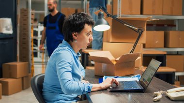 Small business owner preparing merchandise shipment from storehouse shelves and racks, working on products inventory. Young woman planning logistics for quality control supply chain.