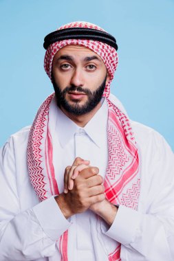 Müslüman adam inanç ve inanç göstererek dua ediyor ve umut dolu bir ifadeyle kameraya bakıyor. Arap duası katlanmış ellerle poz verirken mavi arka planda Tanrı 'nın stüdyo portresini överek
