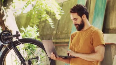 Kırık bisiklet pedalını tamir ederken çevrimiçi tamir rehberleri için dizüstü bilgisayar kullanan beyaz bir erkek. Sağlıklı genç adam mini bilgisayarla internette sörf yaparak bisikletini koruyor..